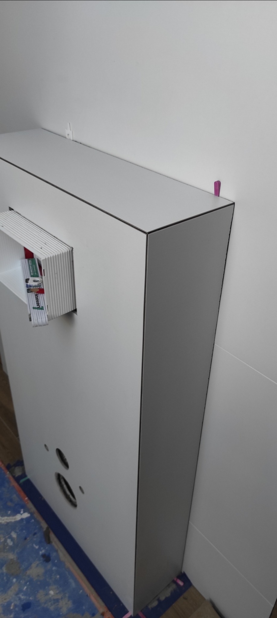 Szara obudowa z otworami i wnęką na narzędzia, zamontowana przy białej ścianie, zabezpieczona taśmą malarską na podłodze.