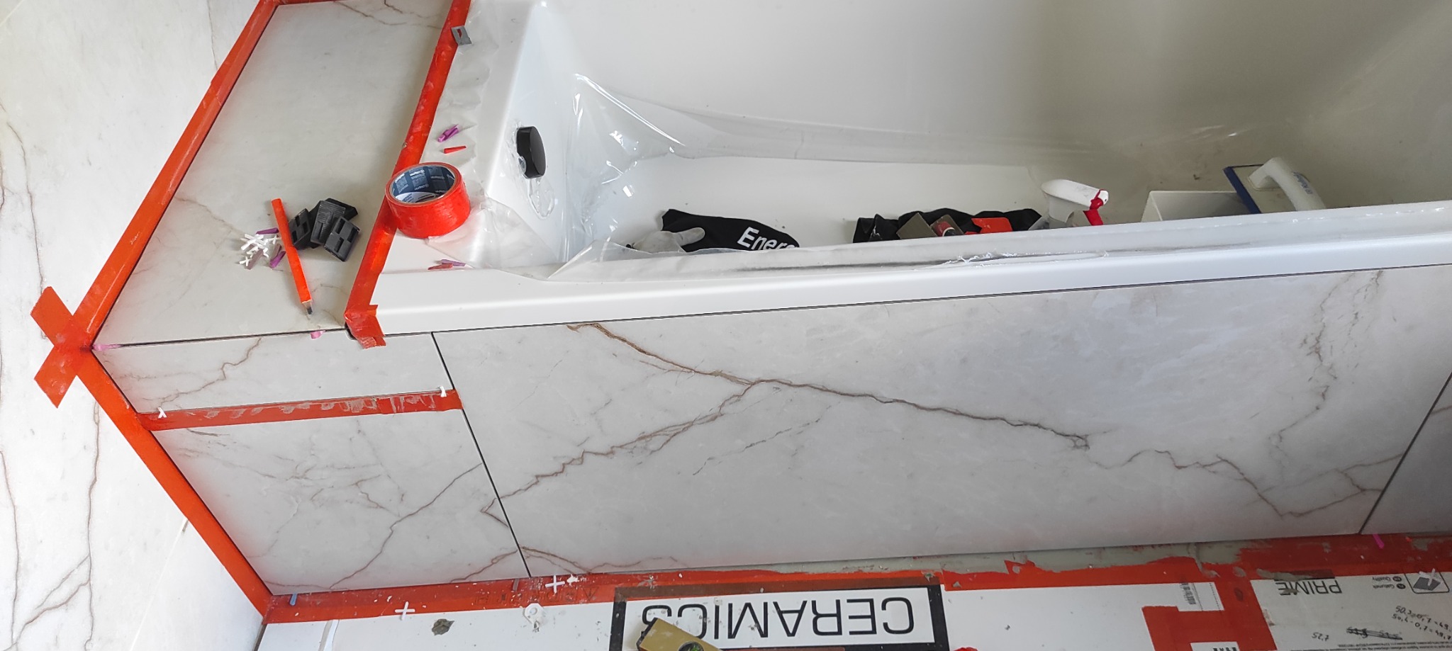Montaż płytek imitujących marmur wokół wanny, z użyciem poziomicy, krzyżyków dystansowych i taśmy malarskiej, widoczne narzędzia w wannie zabezpieczonej folią.