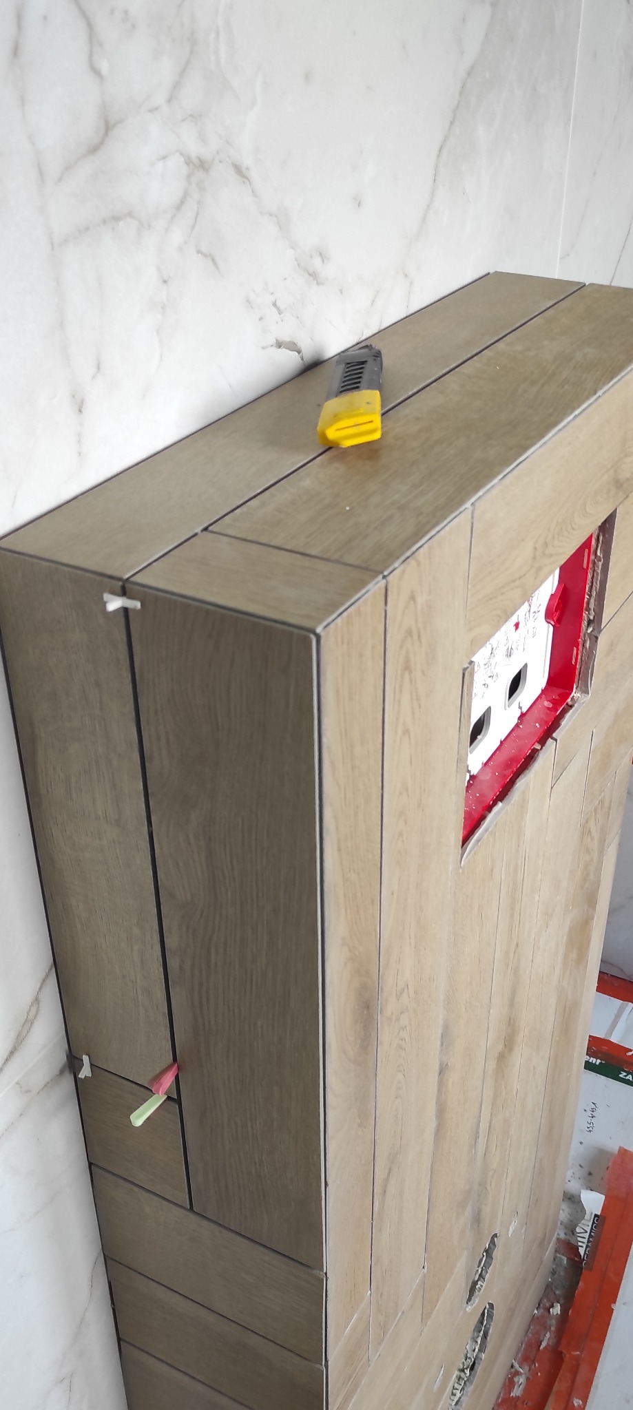 Obudowa stelaża WC wykończona płytkami imitującymi drewno, z widocznymi krzyżykami dystansowymi i nożykiem do cięcia płytek na górze, na tle marmurowej ściany.