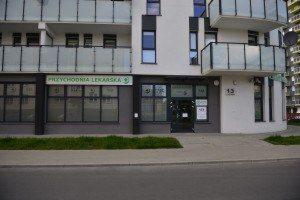 Fasada budynku przychodni lekarskiej z widocznym wejściem, numerem 13 i zielonym logo na szyldzie. Balkony z szklanymi balustradami nad wejściem.