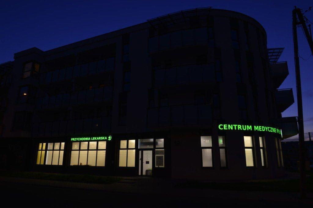 Budynek centrum medycznego z podświetlonymi szyldami 'Przychodnia Lekarska' i 'Centrum Medyczne' o zmierzchu.
