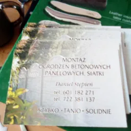 Wizytówka firmy oferującej montaż ogrodzeń betonowych, panelowych i siatki, ułożona na książce o butach, obok sztućców i plamy białej substancji.