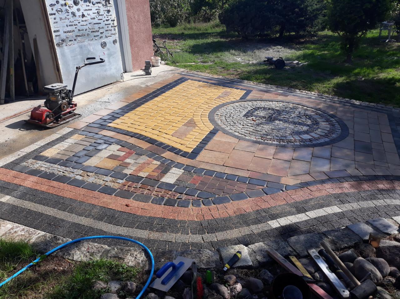 Wzorzysty podjazd z kostki brukowej w różnych kolorach i kształtach, z widoczną zagęszczarką mechaniczną i narzędziami brukarskimi.