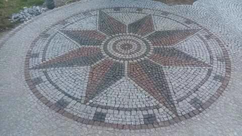 Okrągła nawierzchnia z kostki granitowej ułożona we wzór kompasu z ośmioma ramionami w odcieniach szarości i czerwieni.