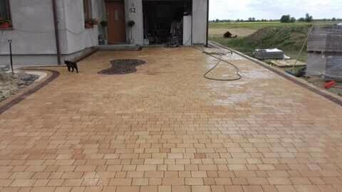 Nowo ułożona, beżowa kostka brukowa przed domem z ciemną obwódką, z widocznym fragmentem dekoracyjnego wzoru oraz czarnym kotem spacerującym po powierzchni.