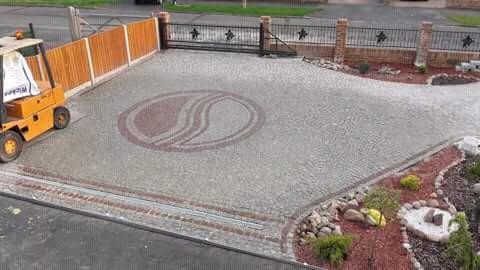 Ułożony z kostki granitowej podjazd z dekoracyjnym wzorem w kształcie yin yang, obok zaparkowany wózek widłowy i rabaty kwiatowe.