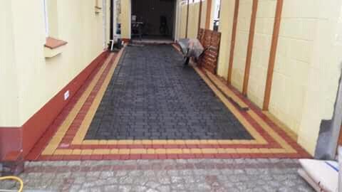 Ułożona, prostokątna ścieżka z ciemnoszarej kostki brukowej z obramowaniem z czerwono-żółtej kostki, prowadząca do garażu, widok z perspektywy