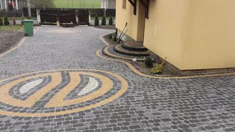 Ułożona szara kostka brukowa z dekoracyjnym wzorem owalnym w kolorze żółtym i białym, prowadząca do wejścia do budynku z trzema kamiennymi stopniami.