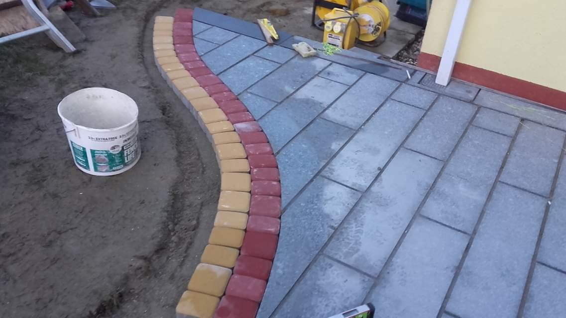 Fragment nowo ułożonej nawierzchni z kostki brukowej w odcieniach szarości, obramowanej łukiem z kostki w kolorach żółtym i czerwonym, z widocznym wiadrem po farbie i narzędziami w tle.