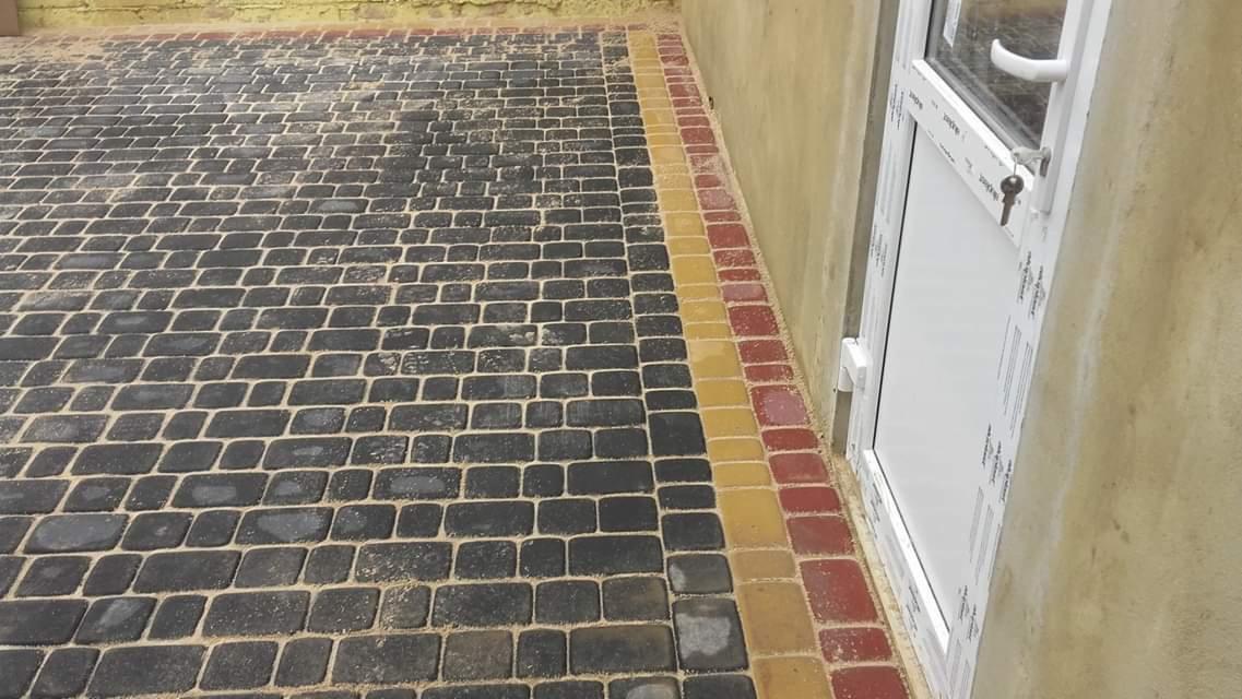 Ułożona kostka brukowa w odcieniach szarości z obramowaniem z kostki czerwonej i żółtej, przylegająca do ściany budynku i białych drzwi.