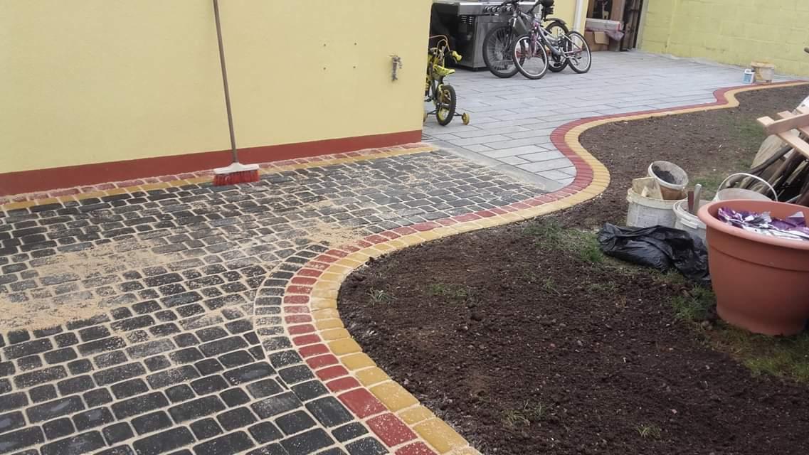 Ułożona kostka brukowa w kolorach czarnym, czerwonym i żółtym, obok przygotowany teren pod trawnik, widoczny budynek z żółtą elewacją i rowerami w tle.