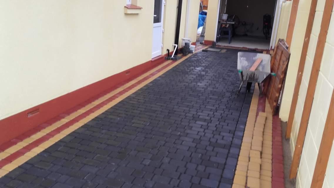 Ułożona ciemnoszara kostka brukowa z obramowaniem w kolorze czerwonym i żółtym, w tle widoczny wózek budowlany i narzędzia.