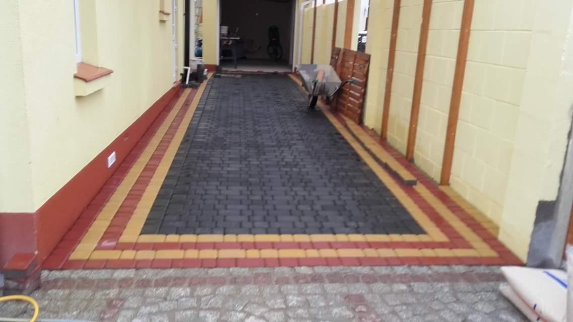 Ułożona, ciemnoszara kostka brukowa z obramowaniem z kostki w kolorach czerwonym i żółtym, prowadząca do garażu, widoczny wózek budowlany i worki z materiałem.