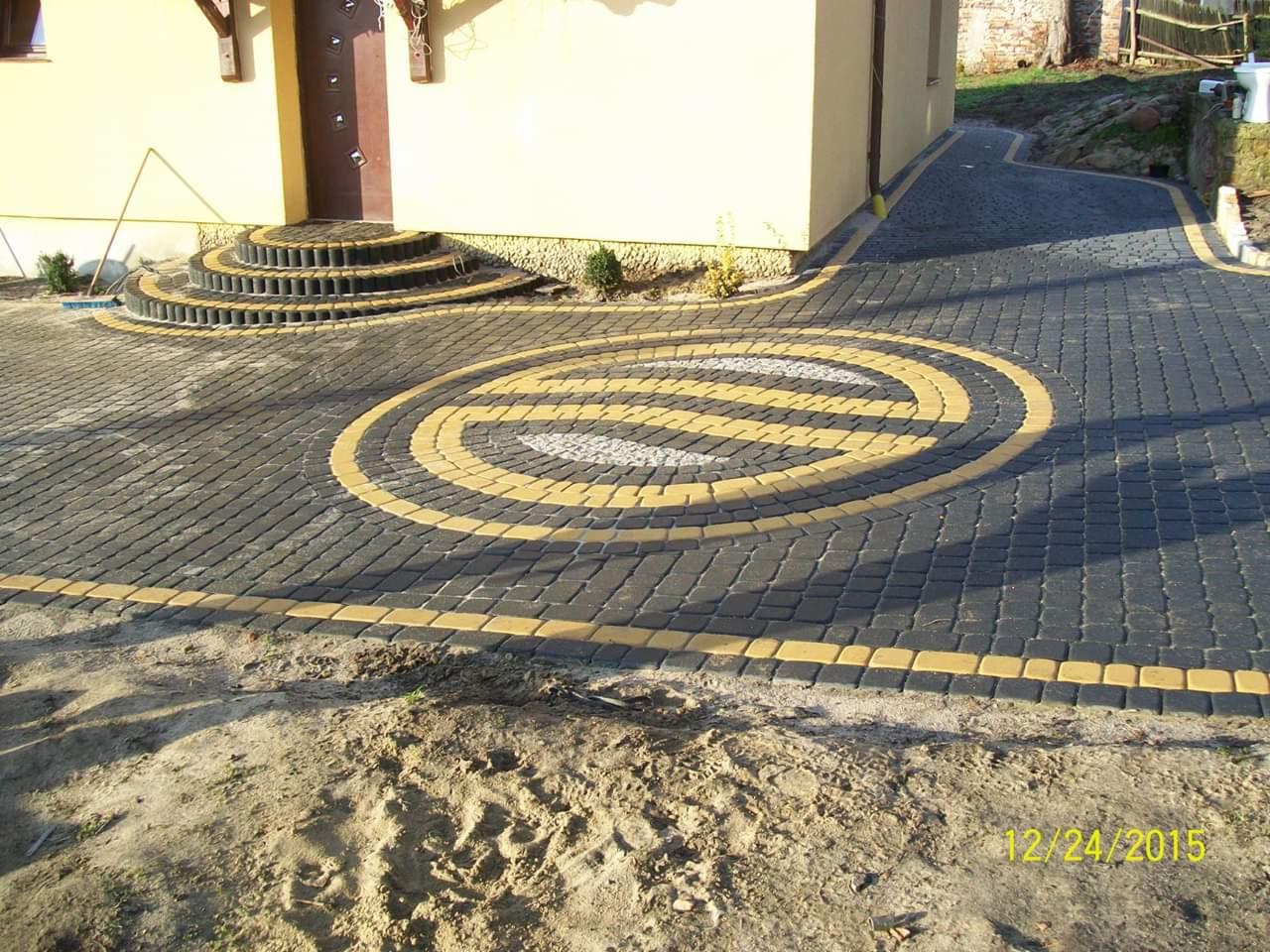 Ułożona kostka brukowa w kolorze grafitowym z żółtymi obramowaniami tworzącymi okrągły wzór przed wejściem do domu z kamiennymi schodami.