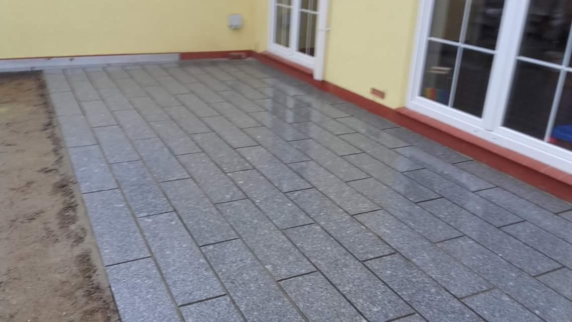 Świeżo ułożona nawierzchnia z prostokątnych płyt granitowych w odcieniach szarości, przylegająca do żółtej ściany budynku z białymi oknami, widoczny fragment przygotowanego podłoża.