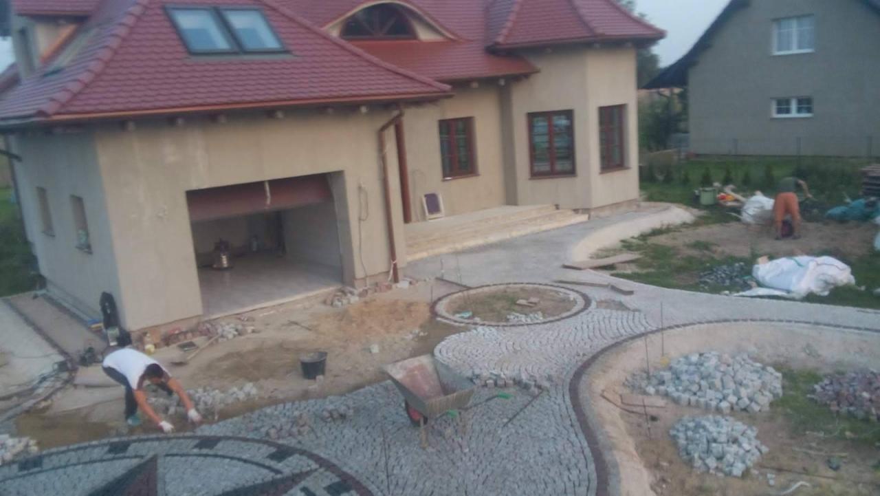 Układanie wzoru z kostki granitowej na podjeździe przed domem; widoczny pracownik w trakcie układania, taczka z narzędziami i materiały budowlane