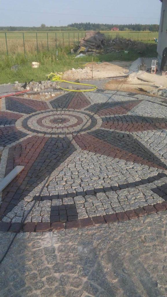 Niedokończona okrągła nawierzchnia z kostki granitowej ułożonej we wzór rozety w odcieniach szarości i czerwieni, widoczny żółty wąż ogrodowy i sterta kostki do ułożenia.
