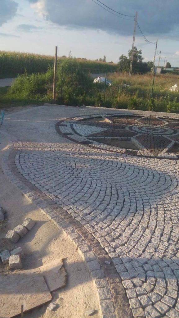 Niedokończona nawierzchnia z kostki granitowej ułożonej w dekoracyjny wzór z widocznymi niedoborami kostki i piaskiem, na tle pola i pochmurnego nieba.