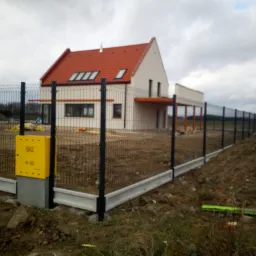 Nowoczesne ogrodzenie panelowe z podmurówką betonową i skrzynką gazową na tle domu jednorodzinnego z czerwoną dachówką.