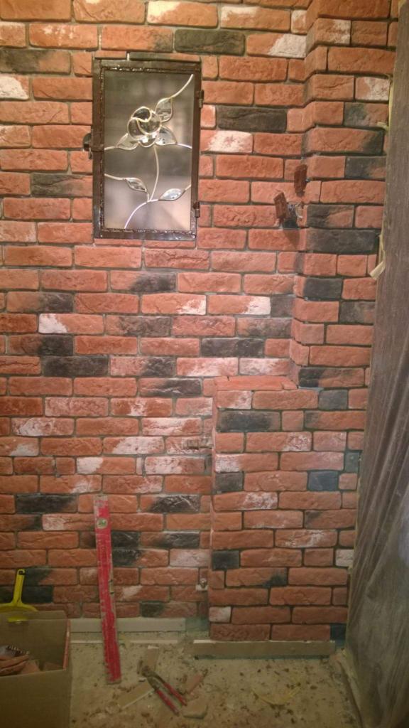 Ściana wyłożona dekoracyjną cegłą w odcieniach czerwieni, bieli i czerni, z wnęką ze szklanym witrażem w kształcie kwiatu, w Leicester. Widoczny poziomica i narzędzia na podłodze sugerują etap prac...