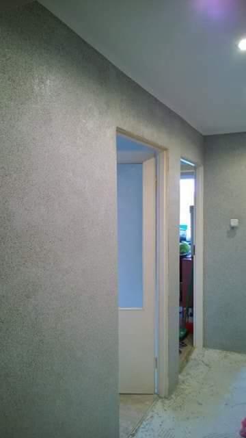 Ściana pokryta szarą, ziarnistą strukturą, widoczne drzwi w białej futrynie i fragment sąsiedniego pomieszczenia, oświetlenie sufitowe.