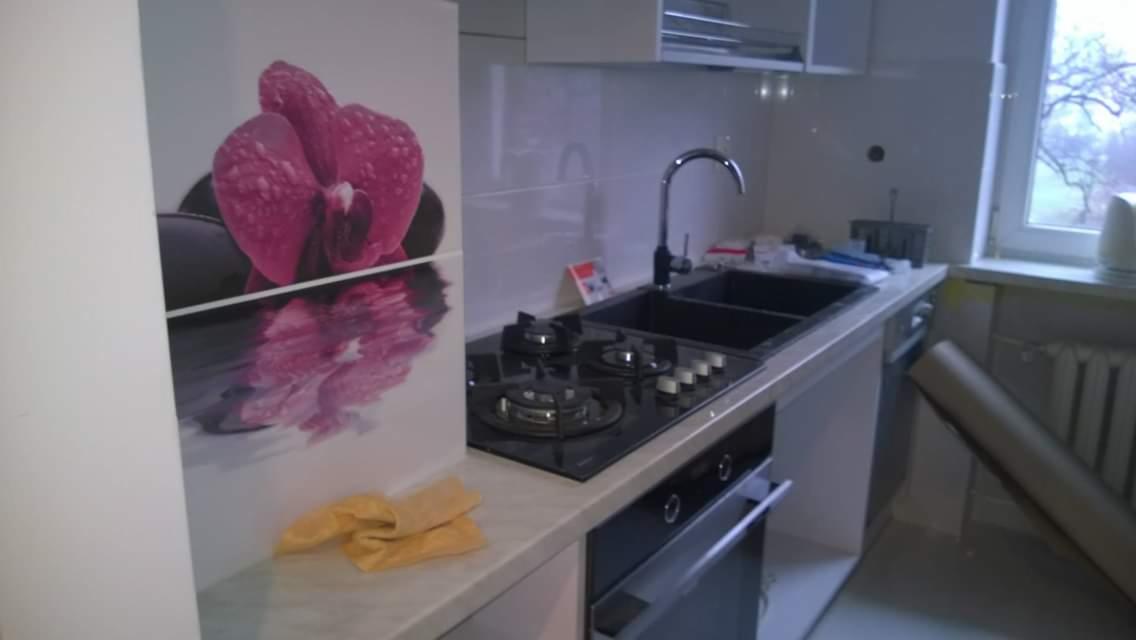 Częściowo wykończona kuchnia z czarnym zlewem, płytą grzewczą i piekarnikiem, na blacie żółta ściereczka, na ścianie panel dekoracyjny z motywem orchidei, widoczne niedoskonałości wykończeniowe...