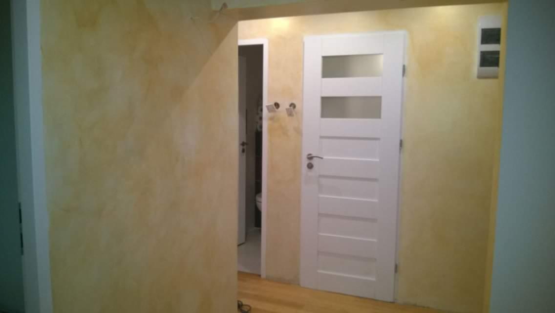 Ściana z efektem dekoracyjnym w odcieniach beżu i żółci, białe drzwi z przeszkleniami prowadzące do łazienki, widoczny fragment instalacji elektrycznej na ścianie.