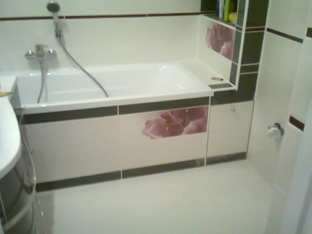 Wykończona łazienka z białą wanną obudowaną panelami z motywem kwiatowym, chromowaną armaturą i białymi płytkami z ciemnobrązowymi akcentami.