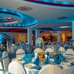 Uroczysta sala bankietowa z okrągłymi stołami nakrytymi białymi obrusami i turkusowymi akcentami, przygotowana na przyjęcie, z dekoracyjnym oświetleniem sufitowym i ozdobną ścianą w tle.