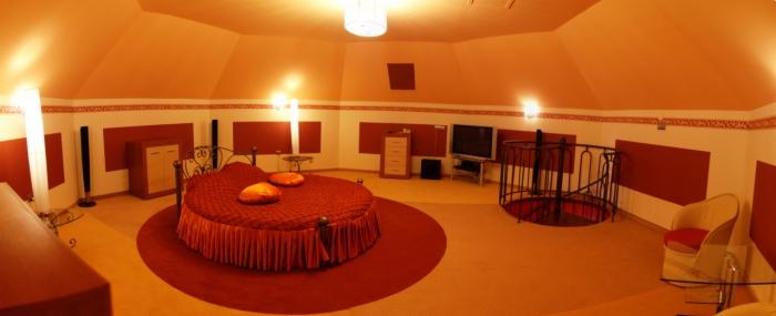 Pokój z okrągłym łóżkiem w kolorze czerwonym, spiralne schody z czarną barierką, pomarańczowe ściany z czerwonymi akcentami i oświetlenie sufitowe.