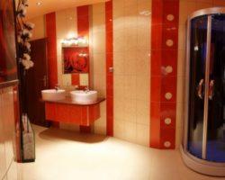 Nowoczesna łazienka z kabiną prysznicową w rogu, umywalkami nablatowymi na czerwonej szafce i pionowymi pasami w kolorze czerwieni i beżu na ścianach.