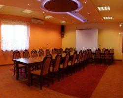 Sala konferencyjna z długim stołem i krzesłami, przygotowana do prezentacji, z rzutnikiem i ekranem w tle.