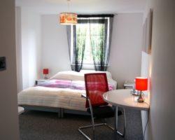 Pokój z łóżkiem, czerwonym krzesłem i okrągłym stolikiem. Lampa z abażurem w geometryczne wzory zwisa nad łóżkiem. Czerwone lampki nocne po obu stronach łóżka. Czarno-białe zasłony w oknie.