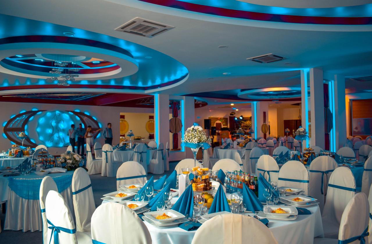 Uroczysta sala bankietowa z okrągłymi stołami nakrytymi białymi obrusami i turkusowymi akcentami, przygotowana na przyjęcie, z dekoracyjnym oświetleniem sufitowym i ozdobną ścianą w tle.