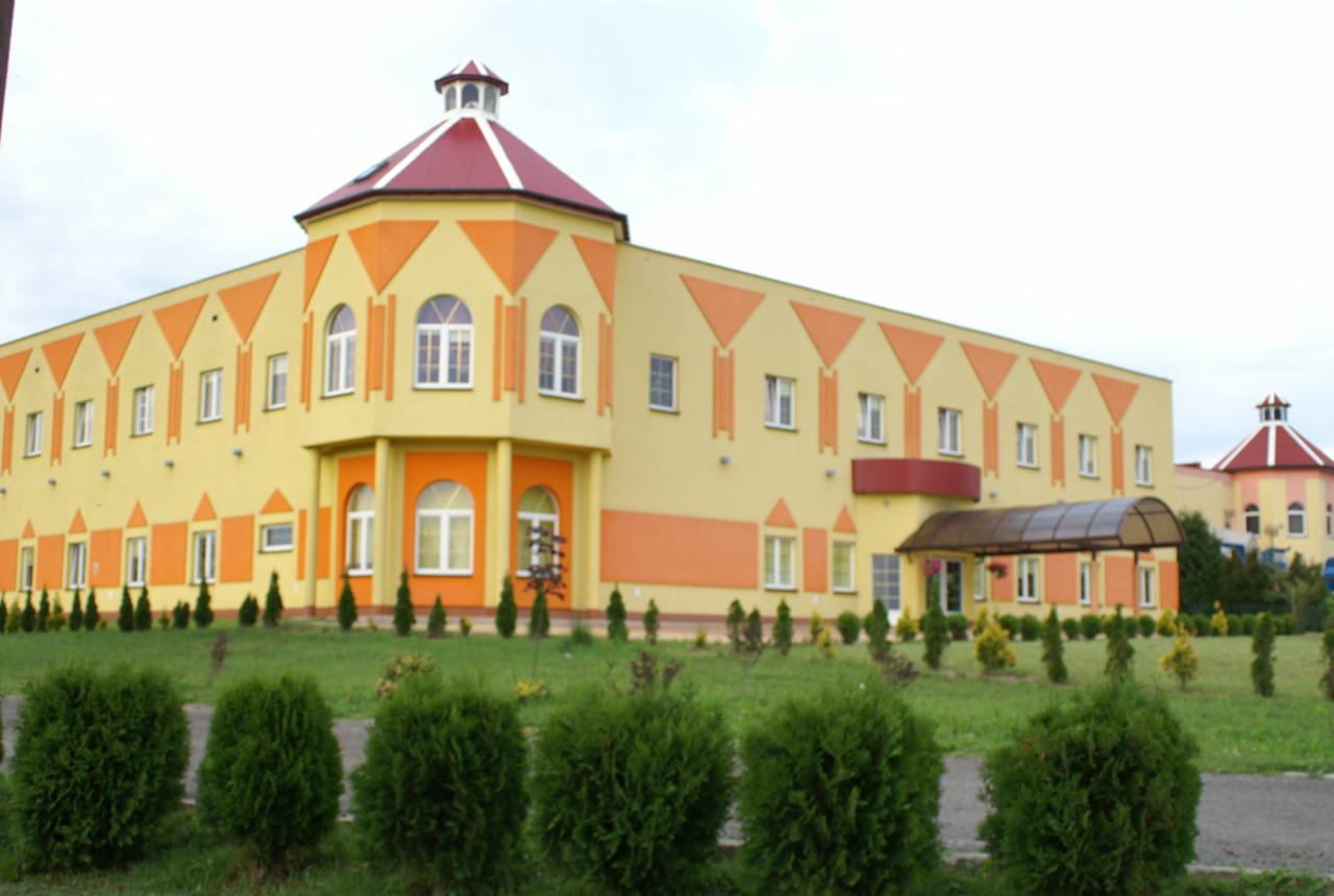 Żółto-pomarańczowy budynek z wieżyczką i czerwonym dachem, otoczony trawnikiem z krzewami.