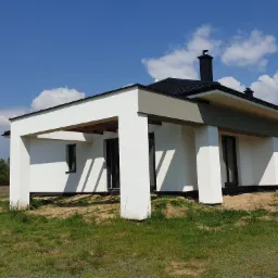 Nowoczesny dom jednorodzinny z płaskim dachem, białymi ścianami i szarymi akcentami, widoczny w słoneczny dzień na tle błękitnego nieba z chmurami.