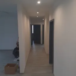 Długi, pusty korytarz po remoncie z szarymi drzwiami i jasnymi ścianami, widoczne ślady po pracach remontowych: karton i folia ochronna przy ścianie.