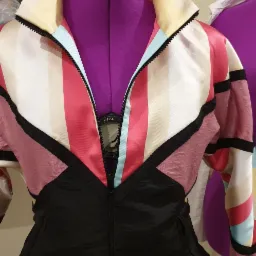 Kolorowa kurtka sportowa w stylu retro z geometrycznymi wzorami na fioletowym manekinie krawieckim, widoczny suwak i kieszenie.