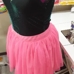 Czarno-zielony top z brokatem i r&oacute;żowa tiulowa sp&oacute;dnica na manekinie krawieckim, obok maszyna do szycia i stosy materiał&oacute;w w tle.