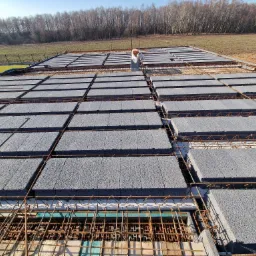 Wykonywanie stropu Teriva z szarego betonu komórkowego zbrojonego stalą, widok z góry na plac budowy z polami i lasem w tle.