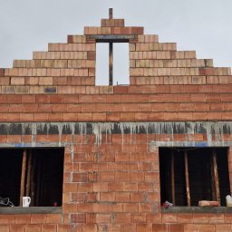 FINISZ USŁUGI REMONTOWO BUDOWLANE SPÓŁKA Z OGRANICZONĄ ODPOWIEDZIALNOŚCIĄ SPÓŁKA Z OGRANICZONĄ ODPOWIEDZIALNOŚCIĄ - Wznoszenie ścian domu z czerwonej cegły ceramicznej Porotherm, widoczne otwory okienne zabezpieczone stemplami, na parapecie czajnik elektryczny i butelka z wodą, w tle pochmurne niebo.