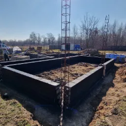 Wykop pod fundamenty z ułożonymi bloczkami betonowymi i zbrojeniem pionowym, widoczny pracownik w tle.