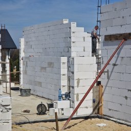 FINISZ USŁUGI REMONTOWO BUDOWLANE SPÓŁKA Z OGRANICZONĄ ODPOWIEDZIALNOŚCIĄ SPÓŁKA Z OGRANICZONĄ ODPOWIEDZIALNOŚCIĄ - Wznoszenie ścian domu z bloczków Ytong w Łodzi; widoczny murarz na rusztowaniu, podpora, bloczki, wiadro, przedłużacz i butelka wody.