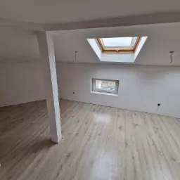 Wnętrze poddasza po remoncie: białe ściany, jasna podłoga laminowana, okna dachowe i okno ścienne, widoczne kable wystające ze ściany, przygotowane do montażu oświetlenia.