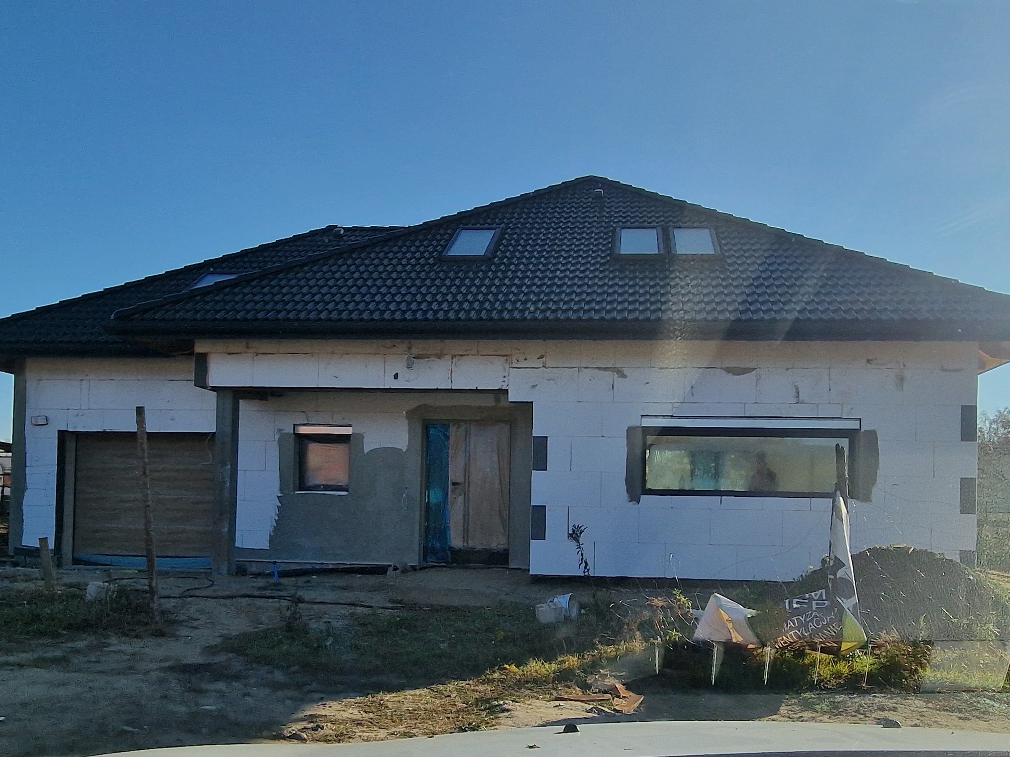 Surowy stan domu jednorodzinnego w trakcie budowy, z widocznymi białymi bloczkami, czarną dachówką z oknami dachowymi, częściowo otynkowanymi fragmentami ścian i prowizorycznie zabezpieczonymi...