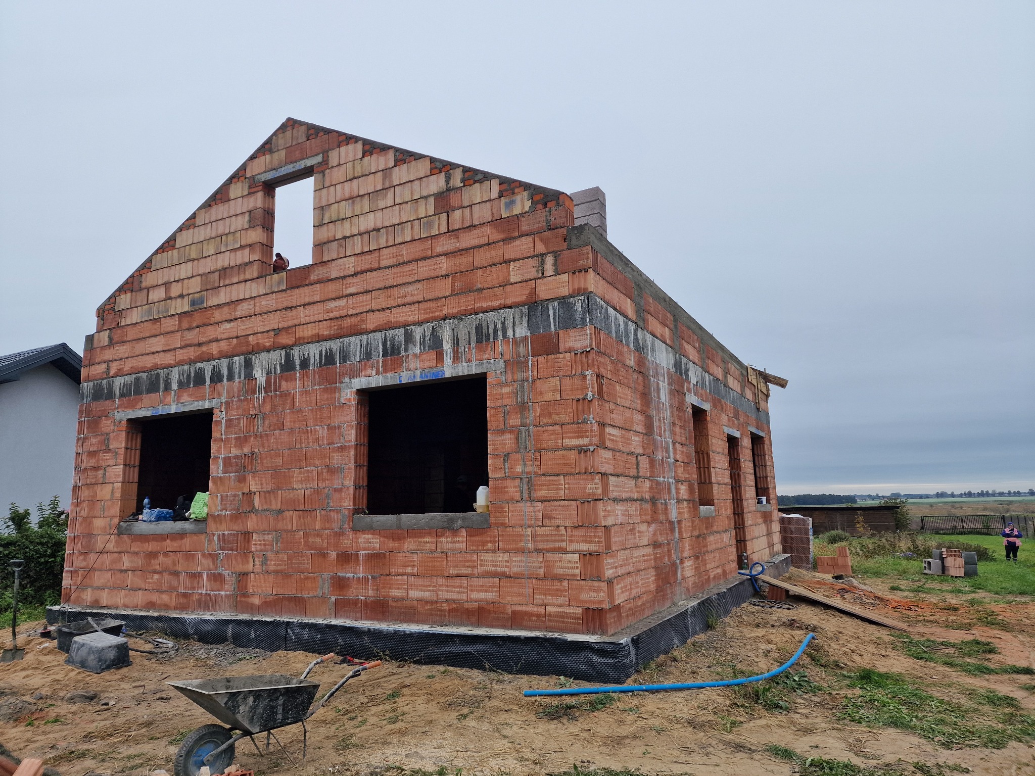 Surowy stan budynku jednorodzinnego z czerwonej cegły, widoczny wózek budowlany, piasek i elementy izolacji fundamentów.