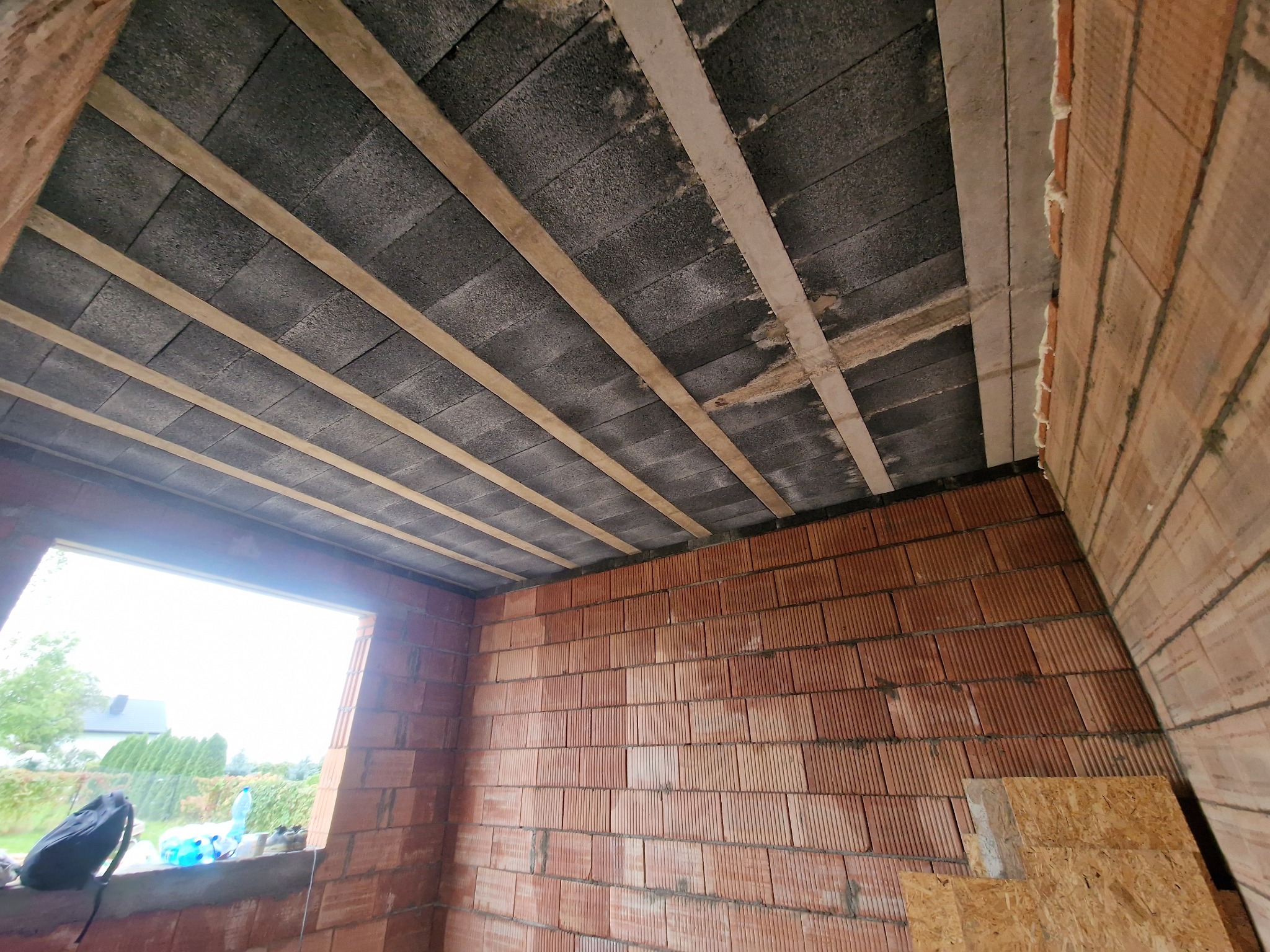 Wnętrze surowego domu w trakcie budowy: ściany z czerwonej cegły, strop z szarych bloczków betonowych wzmocniony drewnianymi belkami, widok przez otwór okienny na zewnątrz.