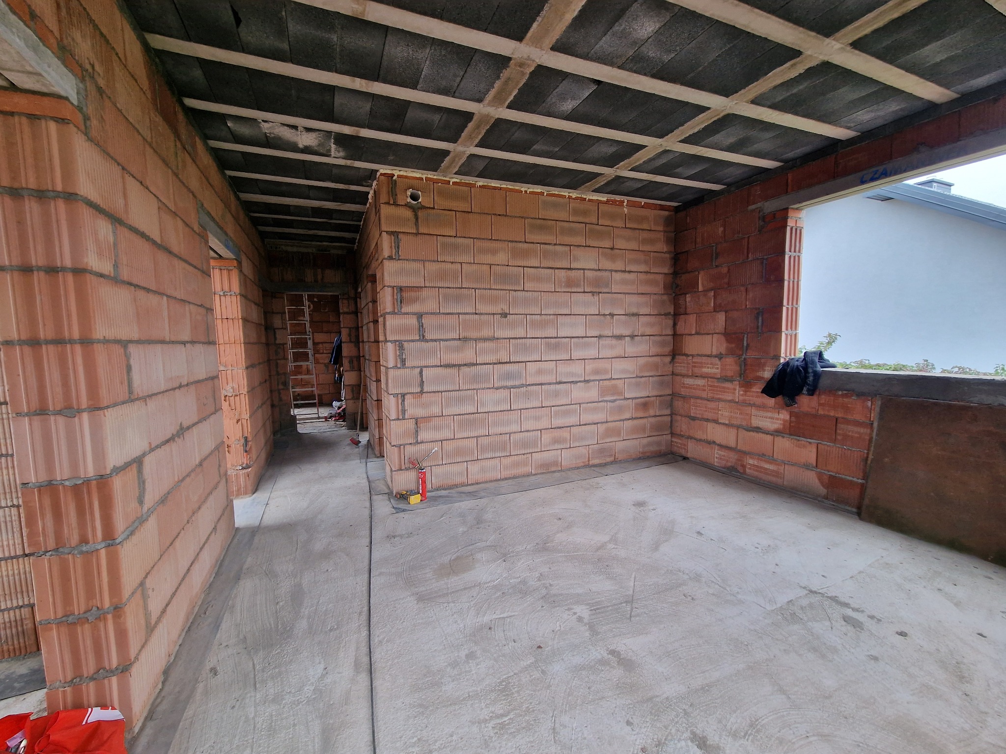 Wnętrze surowego domu w trakcie budowy, widoczne ściany z czerwonej cegły, betonowa podłoga i strop z prefabrykatów betonowych, drabina w głębi pomieszczenia.