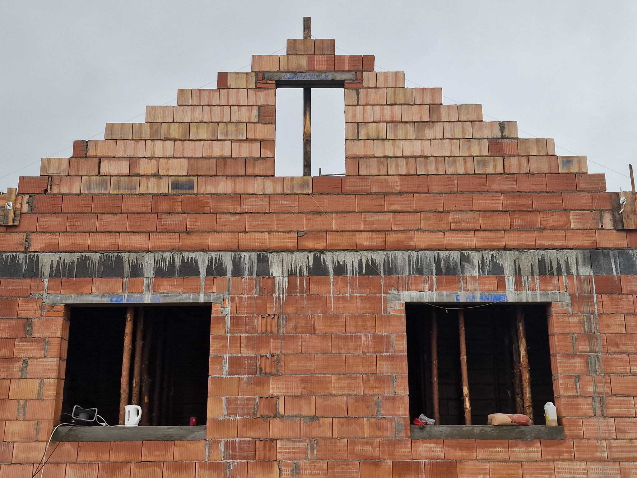 Wznoszenie ścian domu z czerwonej cegły ceramicznej Porotherm, widoczne otwory okienne zabezpieczone stemplami, na parapecie czajnik elektryczny i butelka z wodą, w tle pochmurne niebo.
