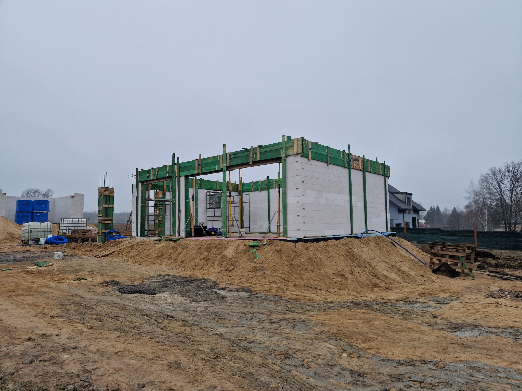 Surowy stan budynku z białych bloczków, z zielonym szalunkiem, na placu budowy z hałdami piasku i widocznymi elementami infrastruktury, w tle zabudowa jednorodzinna i pochmurne niebo.